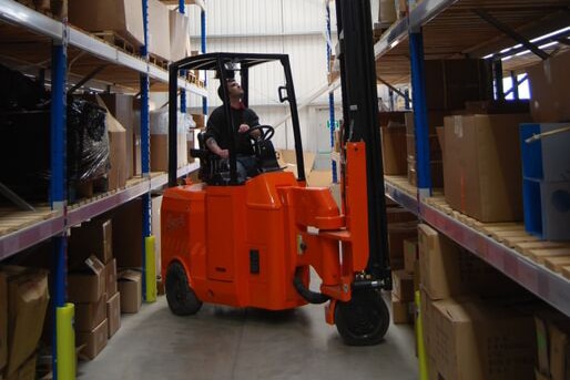 Hombre operando una grúa reach naranja en un almacén, moviendo cajas entre estanterías llenas de productos.