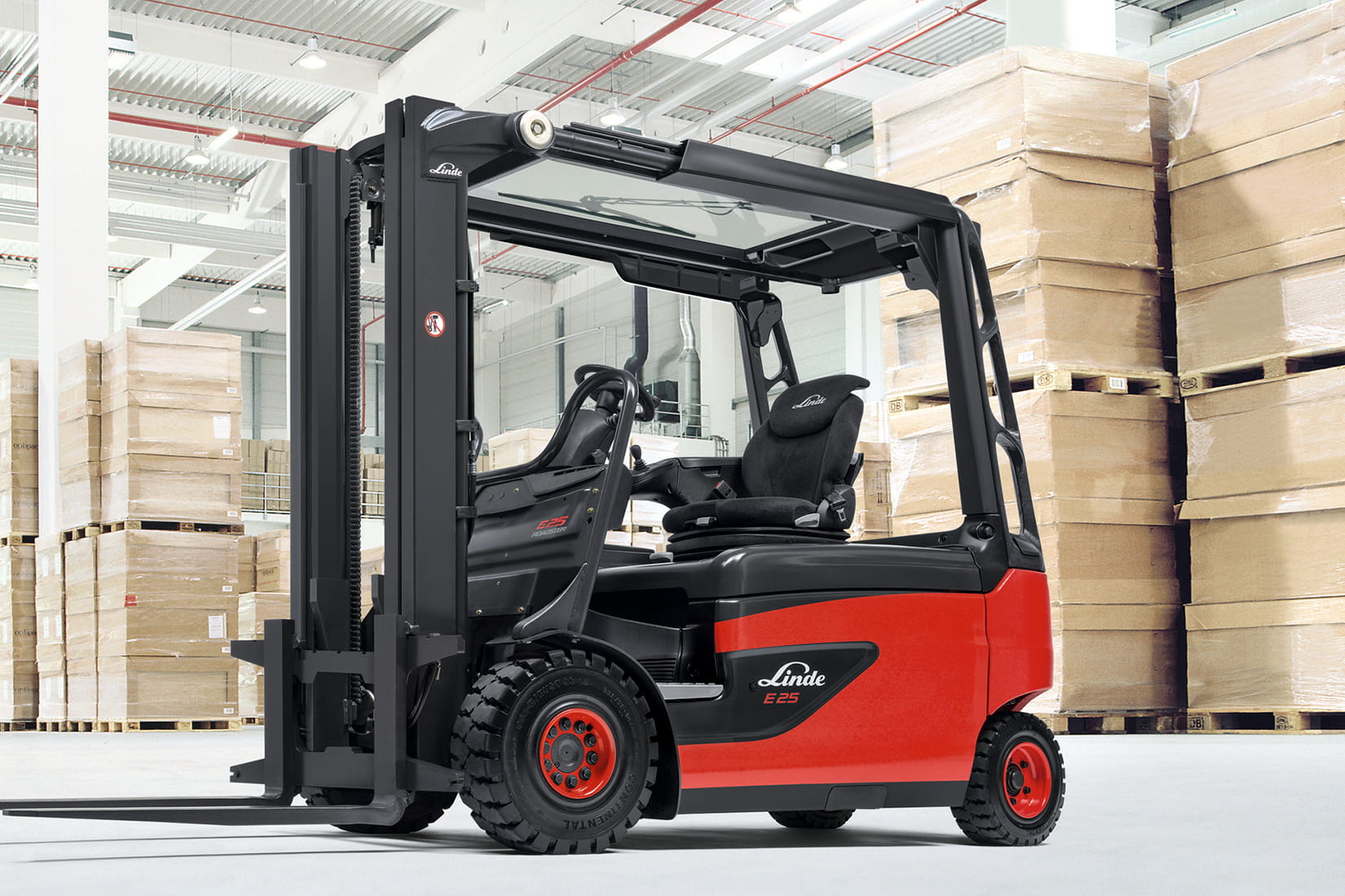 Montacargas eléctrico Linde E25 rojo y negro estacionado en un almacén con cajas apiladas sobre pallets.