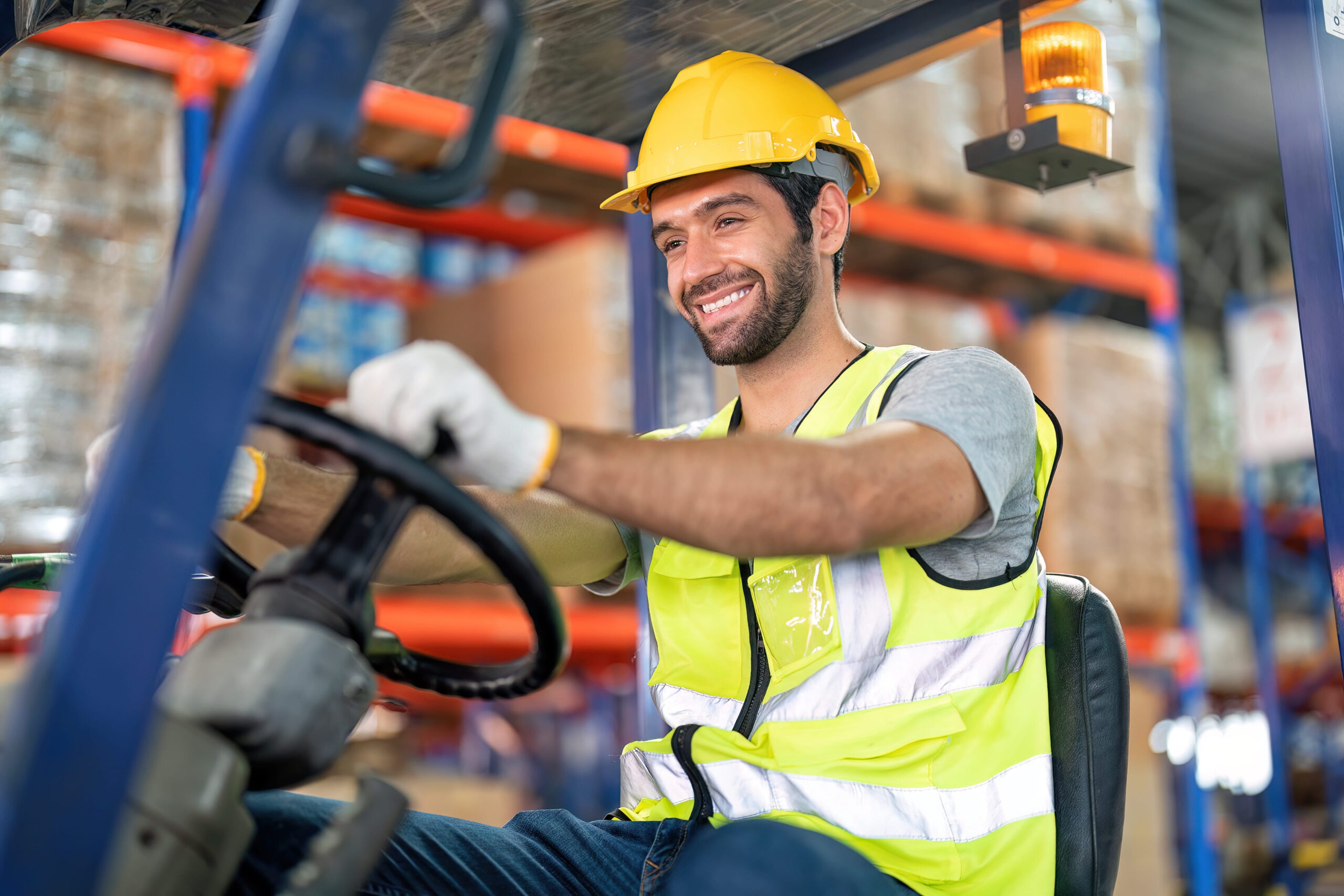 Trabajador con casco amarillo y chaleco reflectante conduciendo un montacargas en un almacén sonriendo.